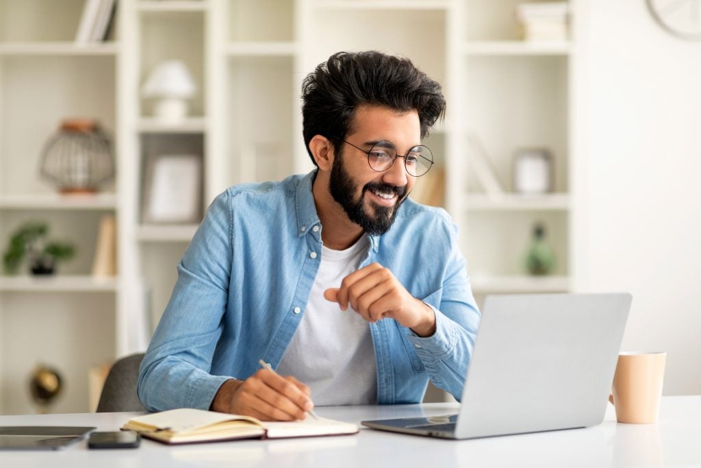 Online Education. Young Indian Man Watching Webinar On Laptop And Taking Notes